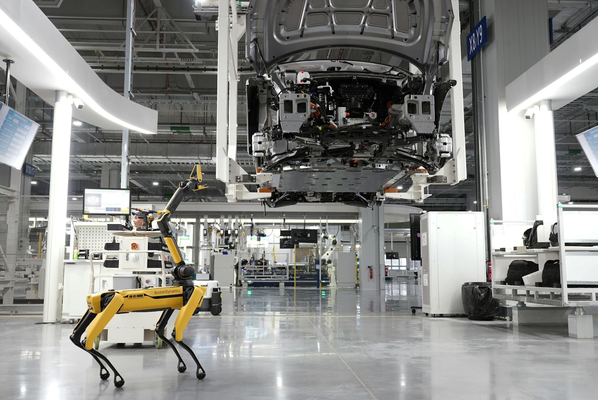 A picture of an electric car on a production line, the car is only half assembled, the factory is white, very new looking and clean. Scattered about the walls there can be seen an abundance of fire extinguishers, these are too small to attempt to tackle a lithium fire with but should suffice if you need to tackle a small fire or, need a melee weapon to take down the small yellow and black Boston Dynamics robot dog that could go rogue at any minute, the dog is located in the foreground of the picture and has a camera or sensor pointing at the half assembled car.