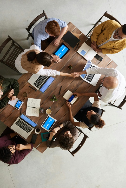 A top down photo of a team around a large rectangle wooden table, there are various laptop computers, tablet computers and phones on the table. Along with the tech there are coffee cups and small plants in tiny pots, as is the custom. Clearly a deal has been done and a firm handshake is taking place between 2 of the people across the table, the other 6 and eagerly anticipating busting out some high fives and hugs.