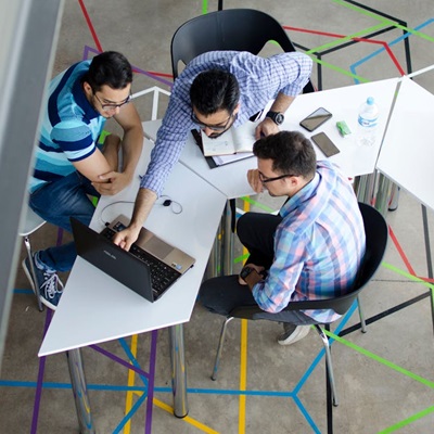 A picture of a classic team around a screen, 3 chaps 1 laptop, an aerial photo looking down on a desk, the floor has one of those crazy carpets with various coloured lines, looks great today but will not age well. While the author of the picture has tried to convince us there are 3 individuals sat at the table no one is being fooled, this is the same person in 3 different positions, right?