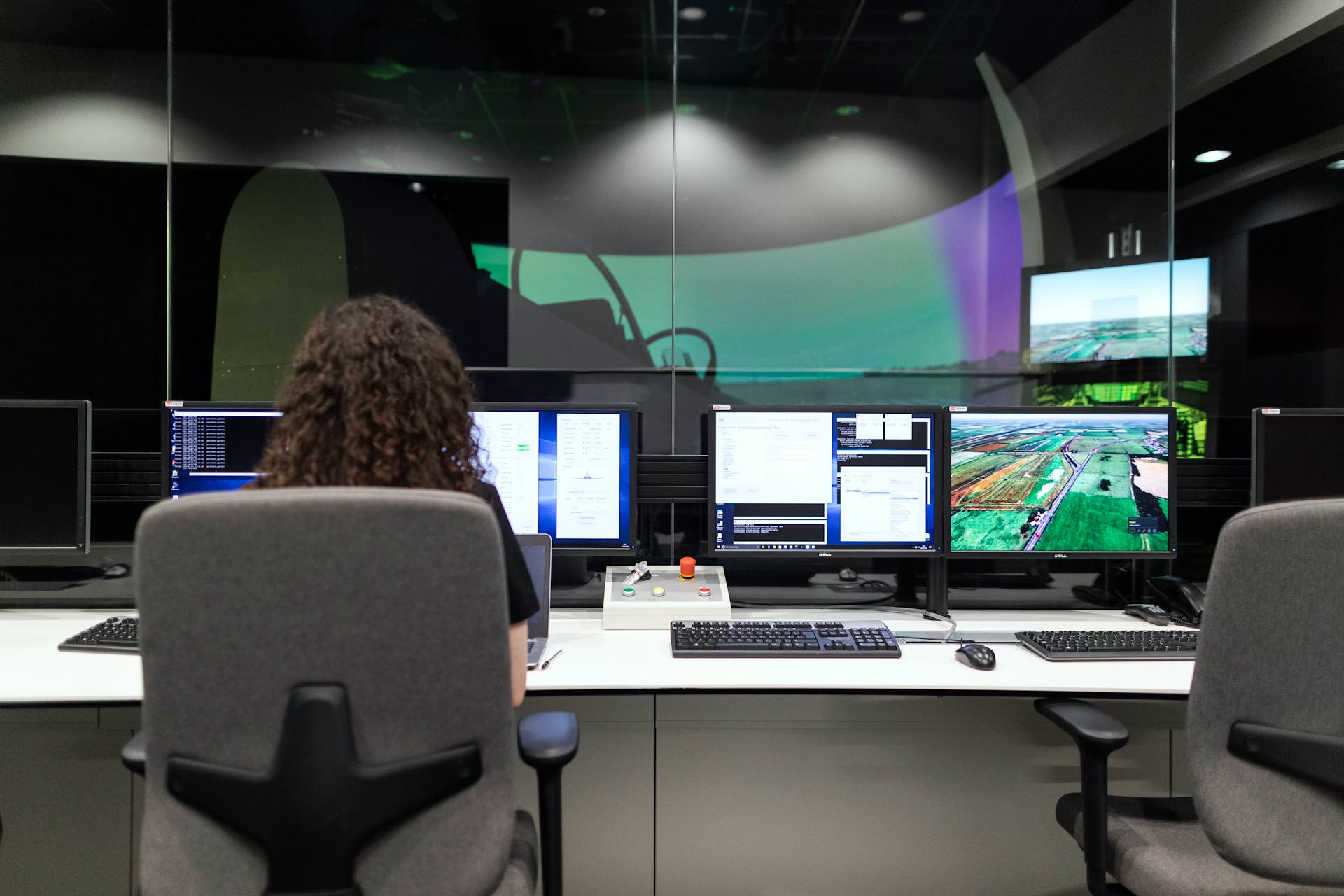 Picture of a curly haired lady sitting at a desk with multiple computers and screens, the lady has her back to the camera, the screens can be clearly seen and show technical information, in from of the desk is a large glass wall and an aircraft simulator can be seen in the distance. The author of the website was looking for some industrial automotive pictures to insert but set a time limit of 3 minutes on himself to find a picture, clearly, he never found what he was looking for in time!